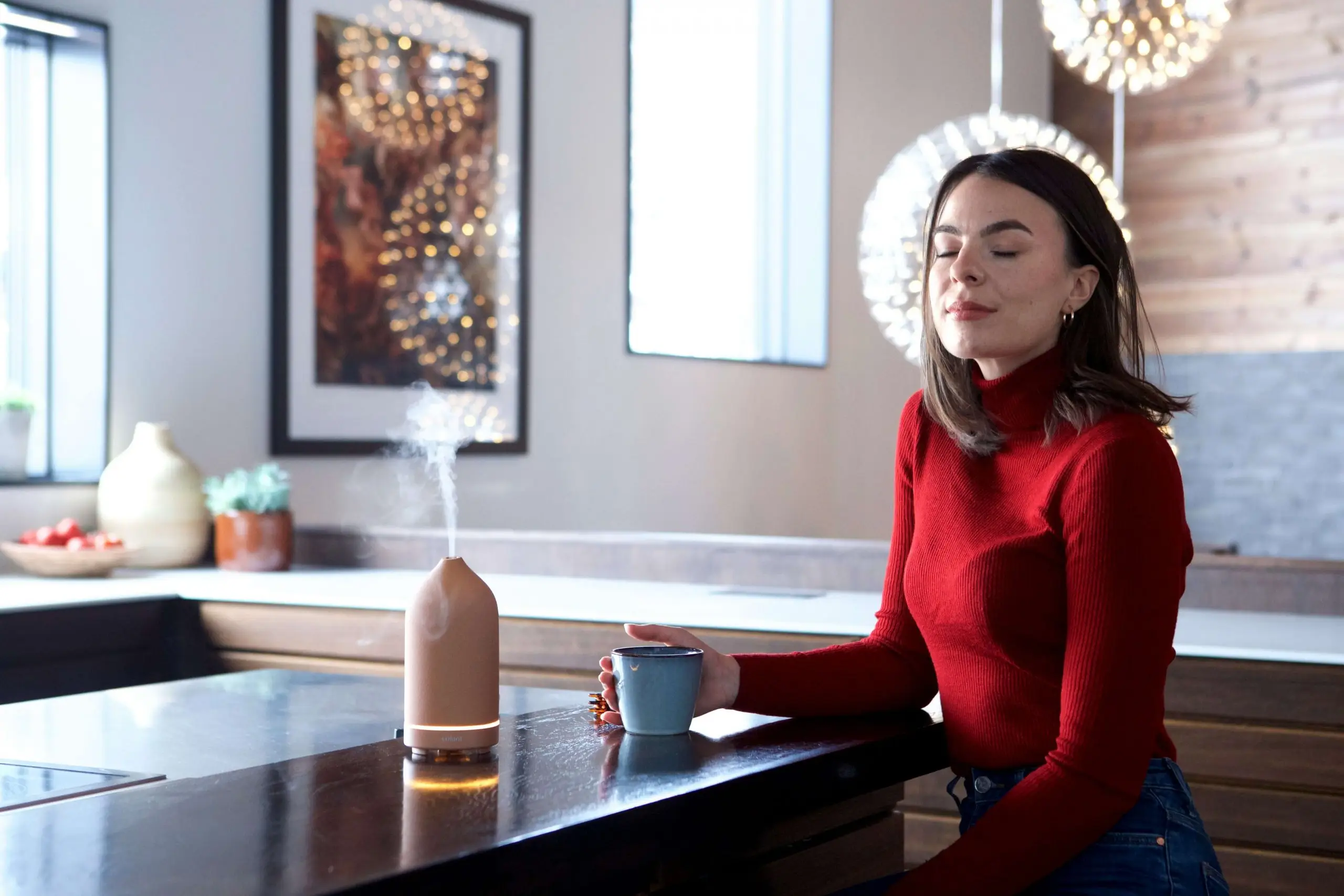 Nainen punaisessa poolopaidassa istuu nykyaikaisessa keittiössä, silmät suljettuina, nauttien kupillisesta juomaa samalla kun diffuusori tuottaa rentouttavaa höyryä taustalla.